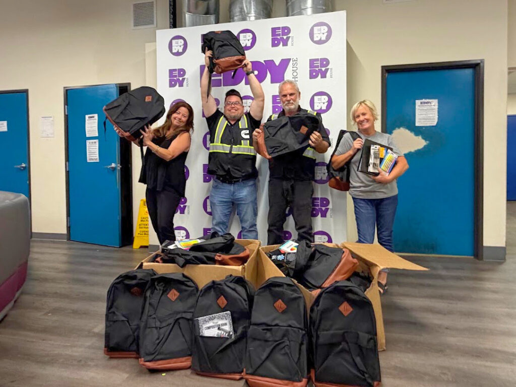 Two Eddy House staff members and two Cummins employees pose with 75 backpacks filled with school supplies, donated through Cummins’ partnership with Foster Love to support young adults served by Eddy House, including youth who have aged out of foster care.