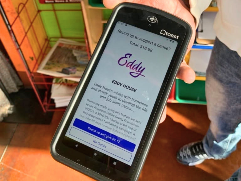 Squeeze In employee holds a point-of-sale terminal displaying a Round Up for Eddy House donation option, inviting customers to add spare change to their purchase in support of homeless and at-risk youth.
