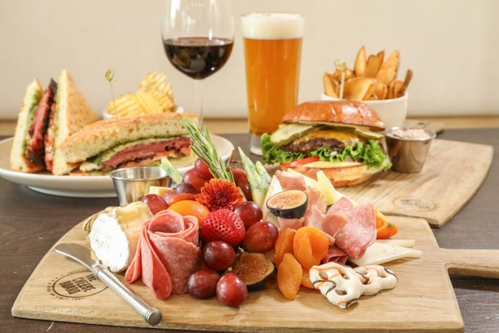 Charcuterie board with cheeses, cured meats, and fruit at The Cheese Board in Reno, with sandwiches, a burger, fries, wine, and beer behind it.